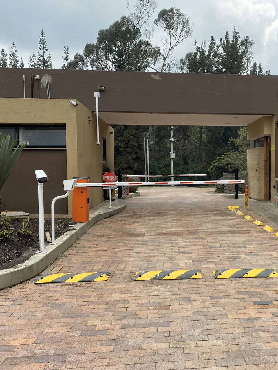 Acceso vehicular con dos talanqueras, columna de control, cámara sobre pedestal apuntando al carril y portería con vista directa sobre la barrera