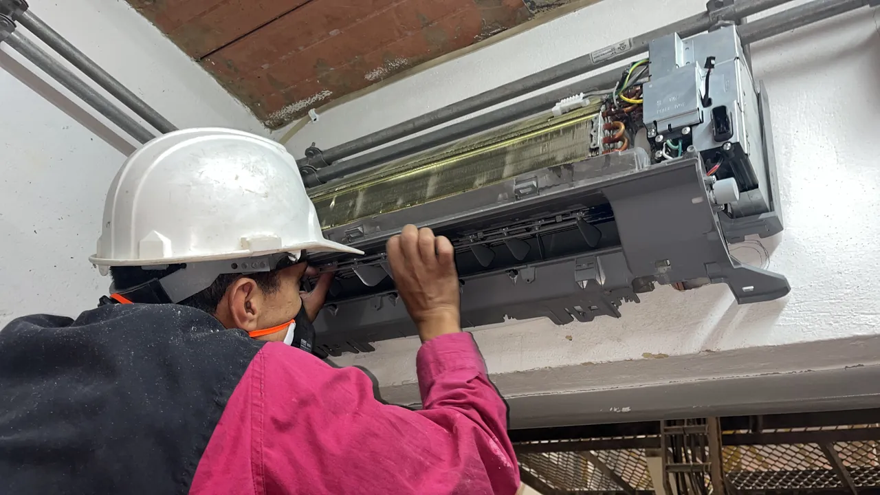 Técnico con casco y tapabocas interviniendo una evaporadora de aire acondicionado abierta, con la tarjeta electrónica a la vista