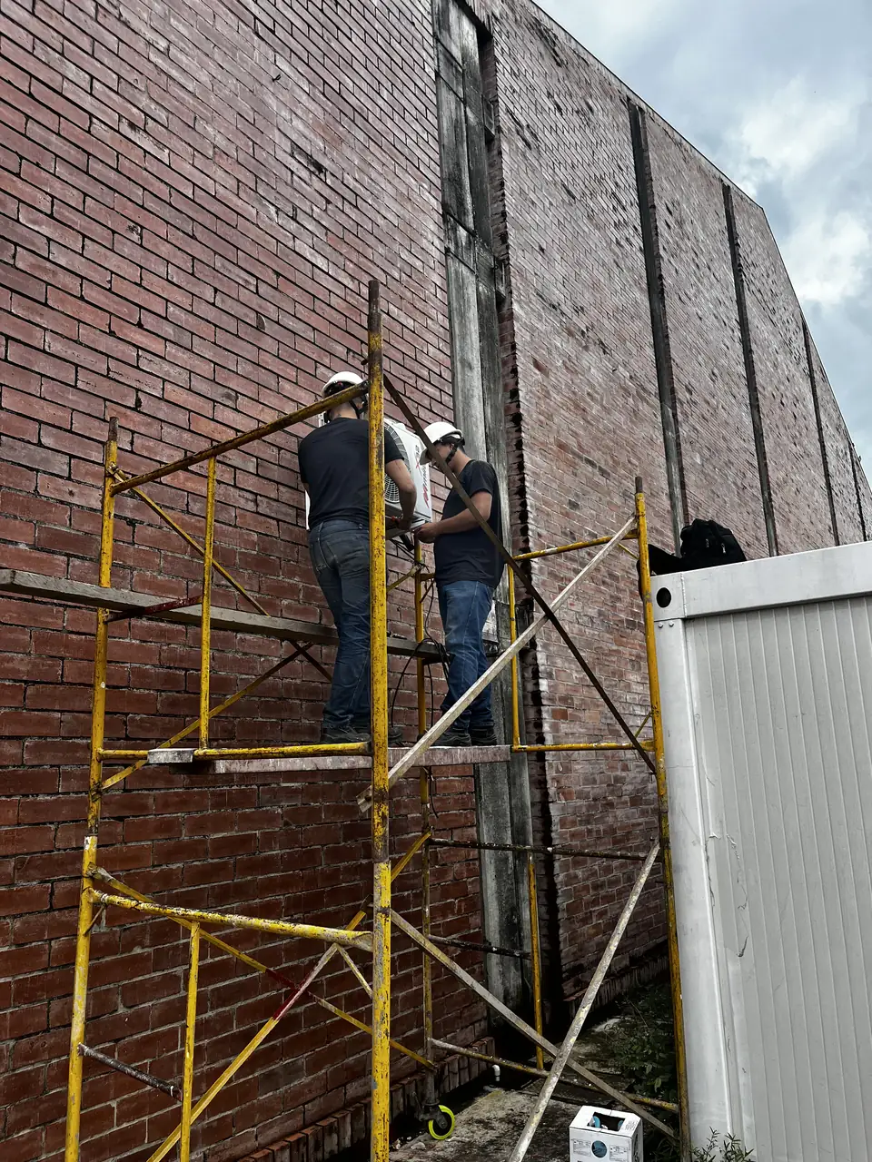 Dos técnicos con casco montando la unidad condensadora de un aire acondicionado sobre la fachada, trabajando desde un andamio