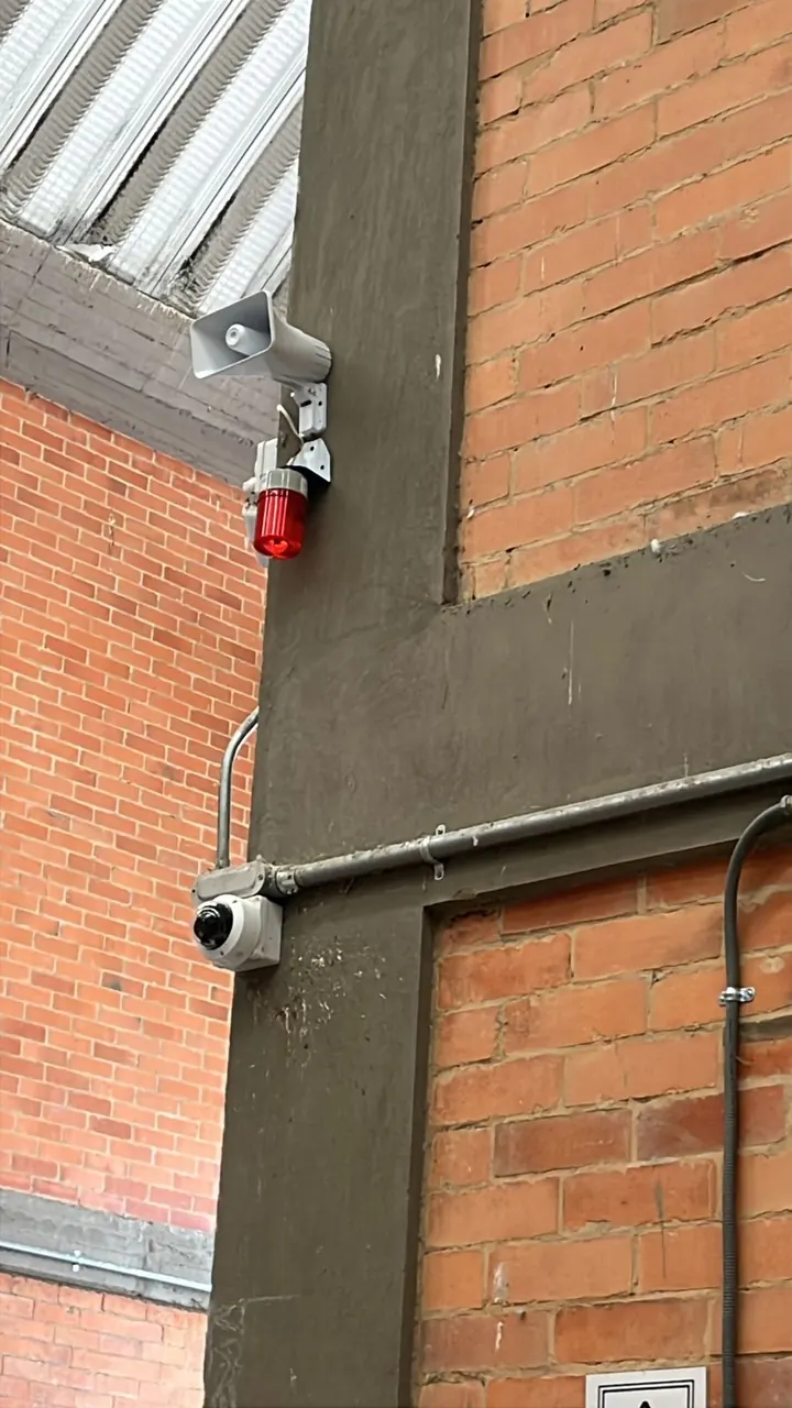 Sirena con luz estroboscópica roja instalada en fachada industrial, junto a una cámara de vigilancia