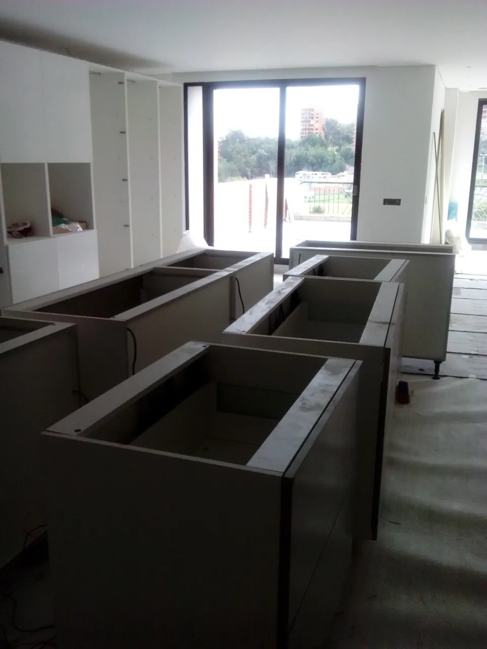 Módulos de mobiliario a medida en pleno montaje dentro de un espacio todavía en obra, con la fachada de ventanal al fondo