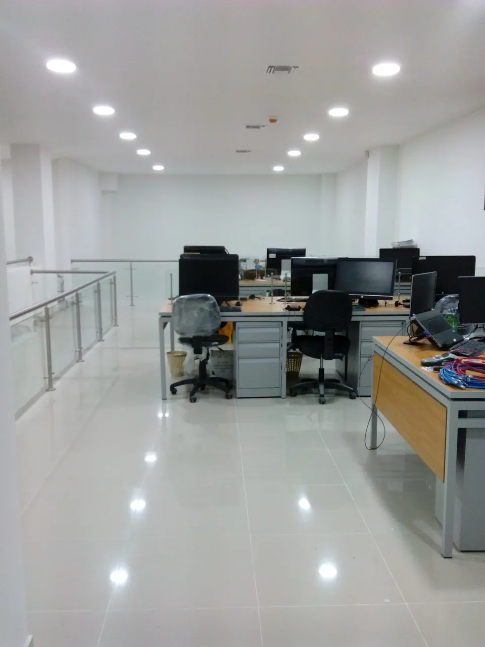 Oficina abierta terminada con piso pulido, iluminación empotrada, puestos de trabajo y baranda de vidrio, con patch cords de red sobre un escritorio