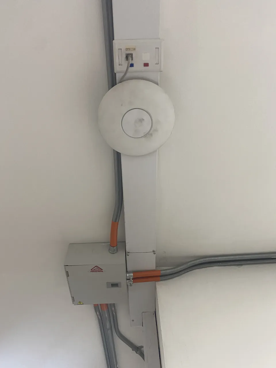 Punto de acceso inalámbrico montado en muro junto a su salida de datos, con canalización metálica a la vista y caja de paso